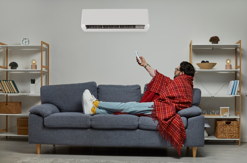 Man on a sofa in New Orleans, trying to keep warm in a cold room with a modern air conditioner, underscoring the importance of efficient HVAC maintenance.