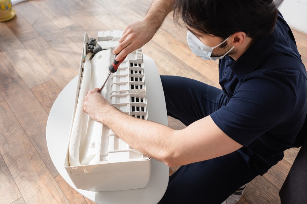 Technician in medical mask fixing a broken air conditioner during HVAC maintenance in New Orleans.