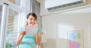 asian woman stand by air conditioner smile happily posing fist gesture about electricity bill saving in hand at home - energy saving concept